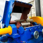 This device is a granulator/shredder, integral to the plastic recycling process. It's designed to break down larger pieces of plastic into smaller granules, facilitating their further processing and recycling. Key features of this machine include a substantial feed hopper that directs materials into the crushing area, alongside a set of visible rotating blades that slice through the plastic. Capable of processing a wide range of plastics, from rigid to flexible types, these machines are essential in plastic recycling facilities. The use of vivid colors on certain parts serves a dual purpose: enhancing safety and providing clear visual cues of the machine's operational components, helping to prevent accidents. Additionally, these machines are typically outfitted with safety mechanisms that automatically halt operations if the machine is opened or accessed in hazardous areas.