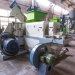 a plastic film squeezer and densifier system. This type of machinery is commonly used in the recycling process to remove excess moisture from plastic films and other flexible plastic materials after washing, and to densify the material to prepare it for further processing steps such as extrusion. The equipment seems to include a hopper for feeding materials into a squeezer or compactor, and a motorized system likely to drive the squeezing process. The setup is typical of a facility that handles large volumes of plastic recycling.