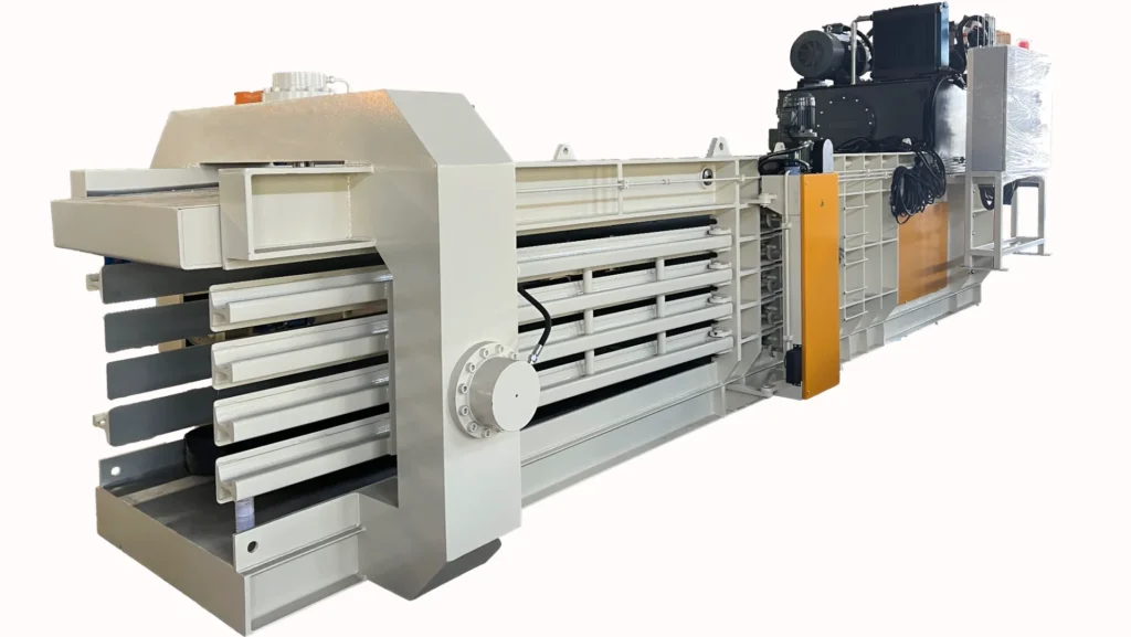 Una empacadora industrial de gran tamaño, diseñada para funcionar de forma totalmente automática con una capacidad de 100 toneladas. Esta maquinaria se utiliza normalmente para comprimir materiales de desecho en pacas compactas, lo que simplifica su manipulación y transporte. La empacadora que se muestra en la imagen es predominantemente gris con detalles en blanco y naranja, e incluye múltiples cámaras de compresión y mecanismos hidráulicos para garantizar un empacado eficiente de los materiales. Este tipo de equipo es crucial en las instalaciones de gestión y reciclaje de residuos para reducir el volumen de residuos y mejorar la eficiencia de los procesos de reciclaje.