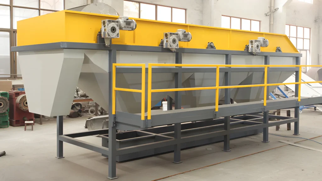 en industriel sink-float separationstank i et produktionsanlæg. Tanken er aflang, med en levende gul top og grå strukturelle komponenter. Flere elektriske motorer monteret på toppen af tanken antyder dens operationelle funktionalitet. Strukturen er understøttet af en robust, mørkegrå metalramme med sikkerhedsgelændere malet i gult. Baggrunden på billedet viser en industriel indstilling med forskellige maskiner og værktøjer spredt rundt. Gulvet er beton, og naturligt lys oplyser rummet gennem vinduer, der er synlige i toppen.